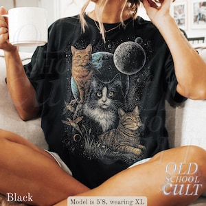 以下が含まれることがあります： 宇宙猫のデザインが施された黒いTシャツ。グラフィックには3匹の猫、惑星、星が含まれています。「OLD SCHOOL CULT」の文字が右下にプリントされています。モデルの身長は173cmで、XLサイズを着用しています。