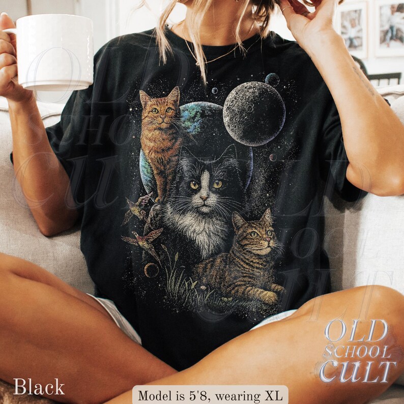 以下が含まれることがあります： 宇宙猫のデザインが施された黒いTシャツ。グラフィックには3匹の猫、惑星、星が含まれています。「OLD SCHOOL CULT」の文字が右下にプリントされています。モデルの身長は173cmで、XLサイズを着用しています。