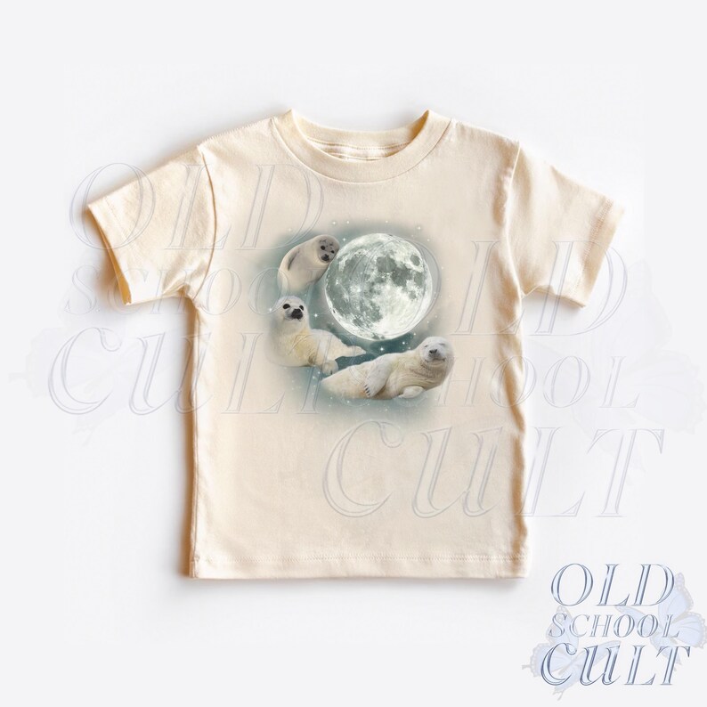 May include: A cream colored t-shirt with a graphic of four baby seals swimming around a full moon. The moon is white and the seals are white with black eyes.