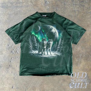 May include: A green t-shirt with a graphic print of two wolves howling at the moon. The wolves are silhouetted against a backdrop of a starry sky and a green aurora borealis.