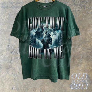 May include: Green t-shirt with a graphic print of two wolves howling at the moon. The text "GOT THAT DOG IN ME" is printed on the shirt.