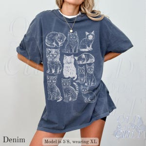 May include: Denim blue t-shirt with a white graphic of various cat illustrations. The shirt has a relaxed fit and short sleeves. The word "Denim" is printed at the bottom left of the image.
