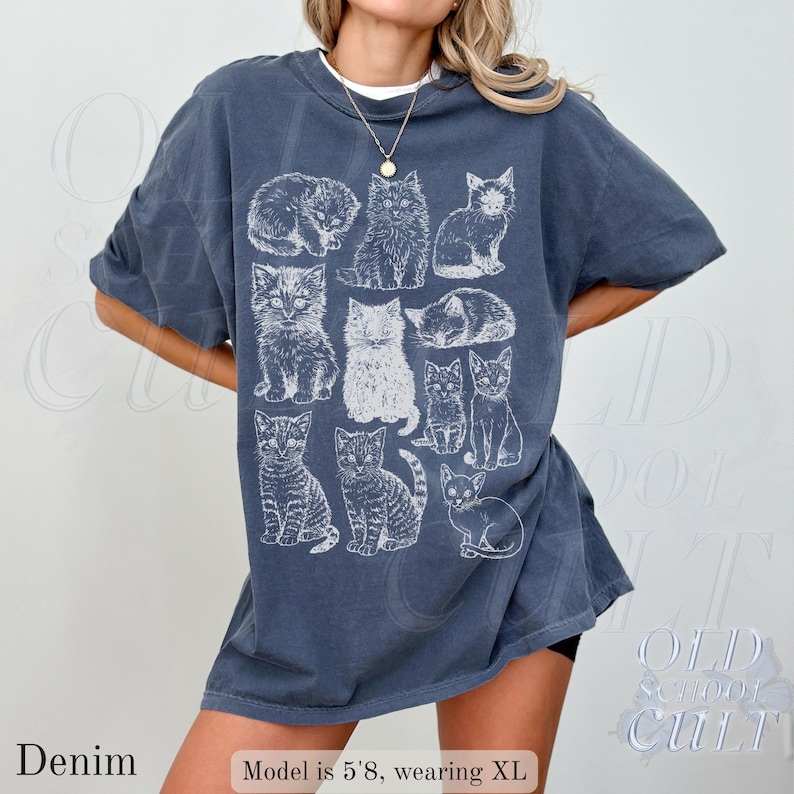 May include: Denim blue t-shirt with a white graphic of various cat illustrations. The shirt has a relaxed fit and short sleeves. The word "Denim" is printed at the bottom left of the image.