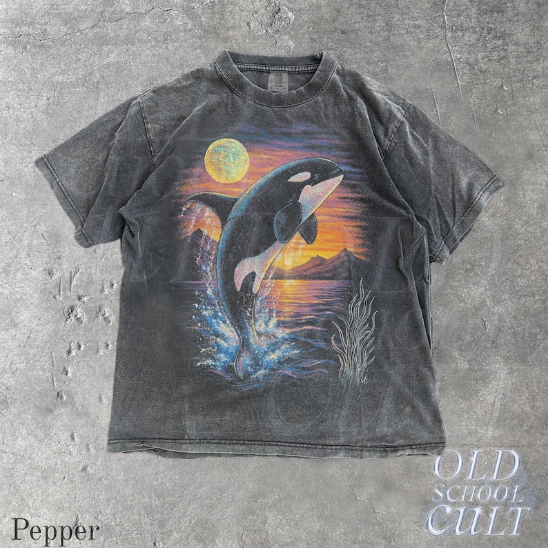 Puede incluir: Camiseta gris oscuro con un gr&aacute;fico de una orca rompiendo la superficie contra un fondo de puesta de sol y luna llena. La camiseta tiene un aspecto vintage y desgastado. El texto "OLD SCHOOL CULT" est&aacute; impreso en la parte inferior derecha.