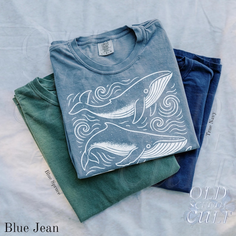 May include: Three t-shirts in blue, green, and navy. The blue shirt features a white graphic of two whales and swirling water. The green shirt is labeled "Blue Spruce" and the navy shirt is labeled "True Navy". The bottom left shirt is labeled "Blue Jean".