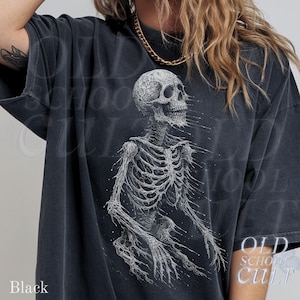 May include: Black t-shirt featuring a detailed white illustration of a human skeleton. The design has a distressed, vintage aesthetic. The text "OLD SCHOOL CULT" is printed on the lower right. The word "Black" is printed on the lower left.