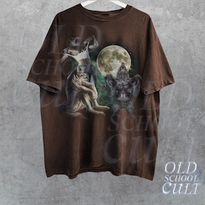 K&ouml;nnte beinhalten: Braunes T-Shirt mit einem Grafikdruck von drei Werw&ouml;lfen, die zum Mond heulen. Die Werw&ouml;lfe sind in einem realistischen Stil mit Fell und scharfen Z&auml;hnen dargestellt. Der Mond ist voll und hell, und der Hintergrund ist ein dunkler Wald.