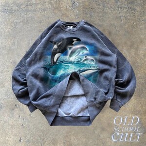 May include: A dark gray sweatshirt with a graphic print of a pod of dolphins and whales jumping out of the water. The sweatshirt has a crew neck and long sleeves.