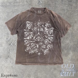 以下が含まれることがあります： エスプレッソ色のTシャツ。フード付きの頭蓋骨が多数描かれた詳細なグラフィックが特徴です。デザインは白とグレーの色合いを使用し、シャツの暗い背景と対照的です。「OLD SCHOOL CULT」の文字が右下に表示されています。