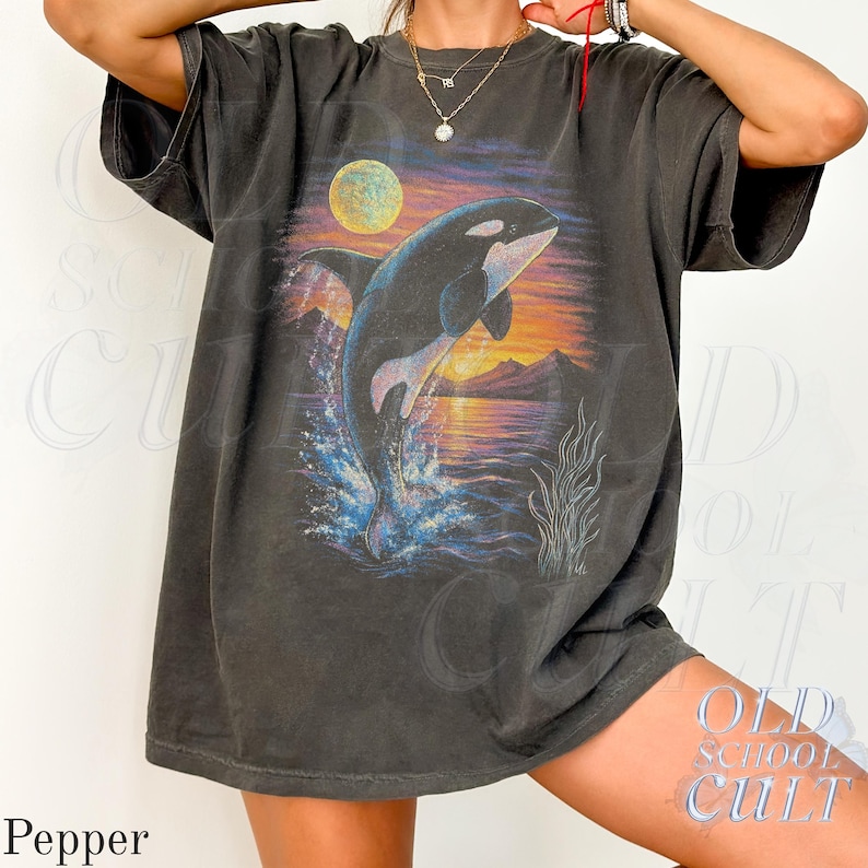 Puede incluir: Camiseta gris oscuro con un gr&aacute;fico de una orca saltando contra un fondo de puesta de sol con luna llena. La camiseta tiene un aspecto vintage y desgastado. El texto "OLD SCHOOL CULT" est&aacute; impreso en la parte inferior derecha.