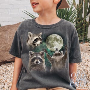 May include: Dark gray t-shirt featuring three raccoons and a full moon. One raccoon is looking up at the moon with its arms raised, while the other two raccoons are looking forward. The text "OLD SCHOOL CULT" is printed on the bottom right.