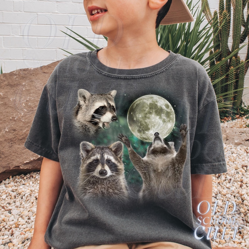May include: Dark gray t-shirt featuring three raccoons and a full moon. One raccoon is looking up at the moon with its arms raised, while the other two raccoons are looking forward. The text "OLD SCHOOL CULT" is printed on the bottom right.
