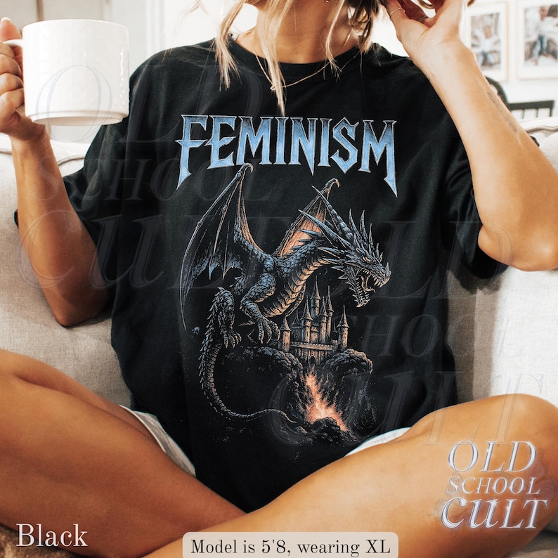 May include: Black t-shirt with the word "FEMINISM" in blue, stylized lettering above a detailed illustration of a dragon perched on a castle. The dragon is black and gray, with a castle in the background. The words "OLD SCHOOL CULT" are in the lower right corner.