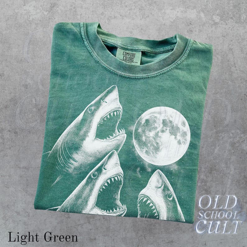 May include: Light green t-shirt featuring a graphic of three sharks with open mouths and a full moon. The shirt has the text "OLD SCHOOL CULT" and "Light Green" printed on it. The shirt is made of a soft material.