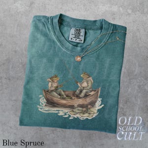 Pode incluir: Camiseta Comfort Colors azul abeto com um gr&aacute;fico de dois sapos pescando em um barco. A camiseta tem gola redonda e uma etiqueta pequena. Um colar de prata com um pingente est&aacute; na camiseta. As palavras "OLD SCHOOL CULT" est&atilde;o no canto inferior direito.