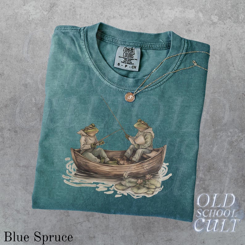 Pode incluir: Camiseta Comfort Colors azul abeto com um gr&aacute;fico de dois sapos pescando em um barco. A camiseta tem gola redonda e uma etiqueta pequena. Um colar de prata com um pingente est&aacute; na camiseta. As palavras "OLD SCHOOL CULT" est&atilde;o no canto inferior direito.