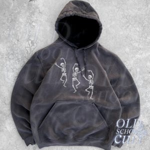 Könnte beinhalten: Ein dunkelgrauer Kapuzenpullover mit Kängurutasche und Kordelzugkapuze. Auf der Vorderseite befindet sich eine weiße Grafik von drei tanzenden Skeletten. Der Hoodie hat einen Vintage-Look.