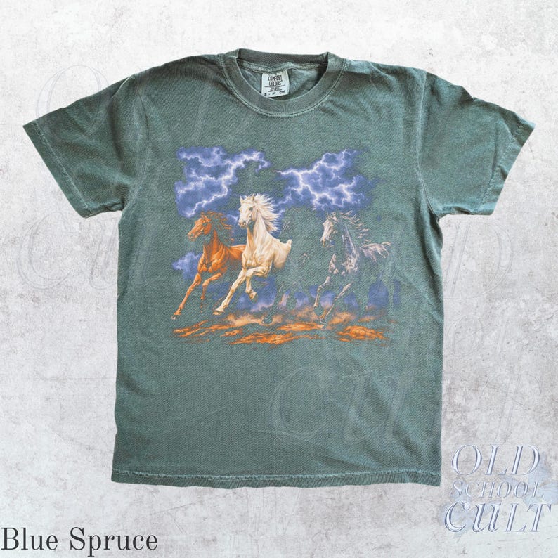 May include: Blue spruce-colored t-shirt featuring a graphic of three running horses with a stormy sky backdrop. The horses are in shades of brown, white, and black. The shirt has a crew neck and short sleeves. The text "Blue Spruce" is at the bottom.