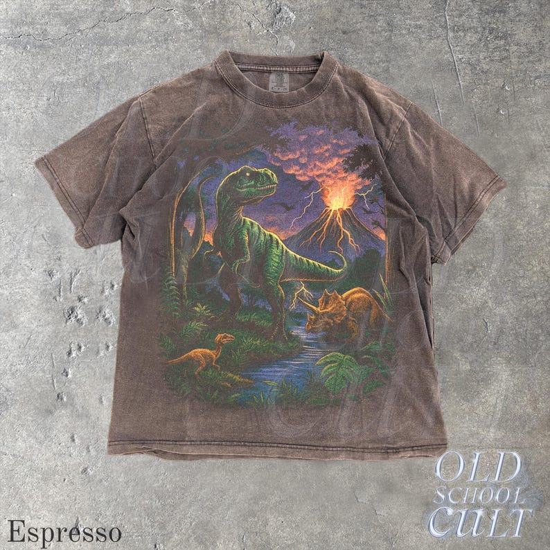 Op de afbeelding: Espresso-kleurig T-shirt met een prehistorische sc&egrave;ne met een groene Tyrannosaurus Rex, een Triceratops en een vulkaan die uitbarst onder een zonsonderganghemel. Het shirt heeft de tekst "OLD SCHOOL CULT" en "Espresso" erop gedrukt.