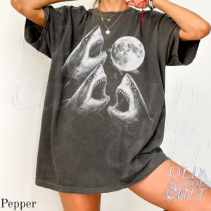 May include: Dark gray t-shirt featuring a graphic of three sharks with open mouths and a full moon. The design is in white, creating a striking contrast against the shirt's color. The shirt has short sleeves and a relaxed fit, suitable for casual wear.