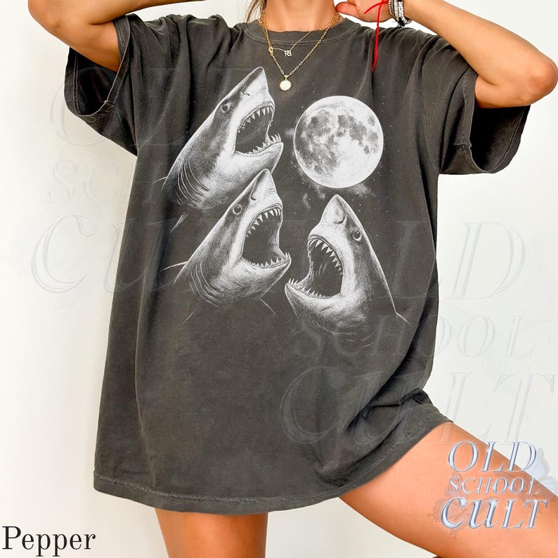 May include: Dark gray t-shirt featuring a graphic of three sharks with open mouths and a full moon. The design is in white, creating a striking contrast against the shirt's color. The shirt has short sleeves and a relaxed fit, suitable for casual wear.