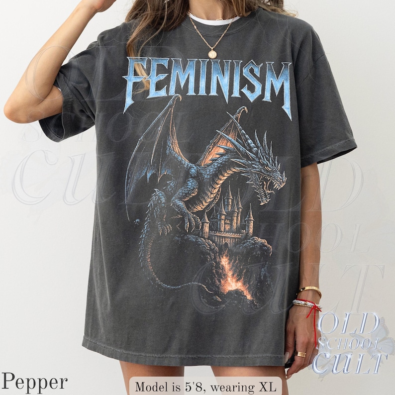 May include: Dark grey t-shirt with the word "FEMINISM" in blue, gothic-style lettering. A detailed illustration of a dragon, castle, and flames is beneath the text. The shirt is a relaxed fit.