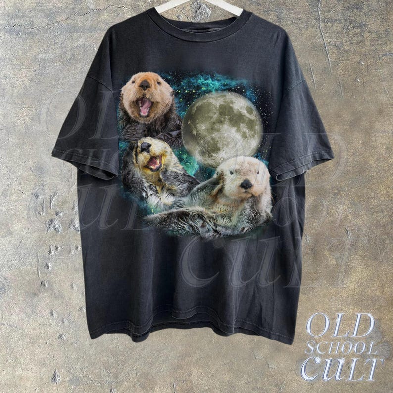 May include: A black t-shirt with a graphic print of three sea otters swimming in front of a full moon. The otters are surrounded by a blue and green nebula-like background.