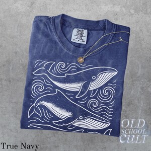 May include: A navy blue t-shirt with a white whale graphic and the text "Old School Cult." The shirt has a crew neck and short sleeves. A gold necklace with a pendant is draped over the shirt. The shirt is labeled "True Navy."
