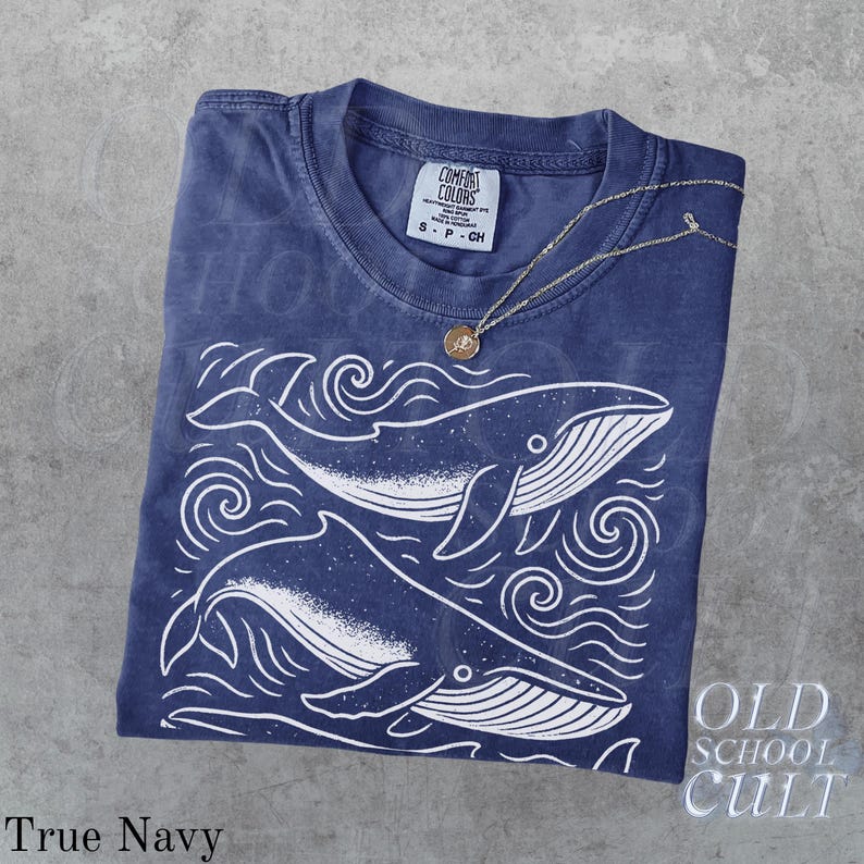 May include: A navy blue t-shirt with a white whale graphic and the text "Old School Cult." The shirt has a crew neck and short sleeves. A gold necklace with a pendant is draped over the shirt. The shirt is labeled "True Navy."