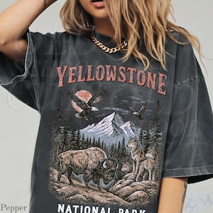 May include: Dark gray t-shirt with a vintage-style graphic featuring the text "YELLOWSTONE NATIONAL PARK." The design includes eagles, mountains, a bison, and wolves, evoking a national park theme.