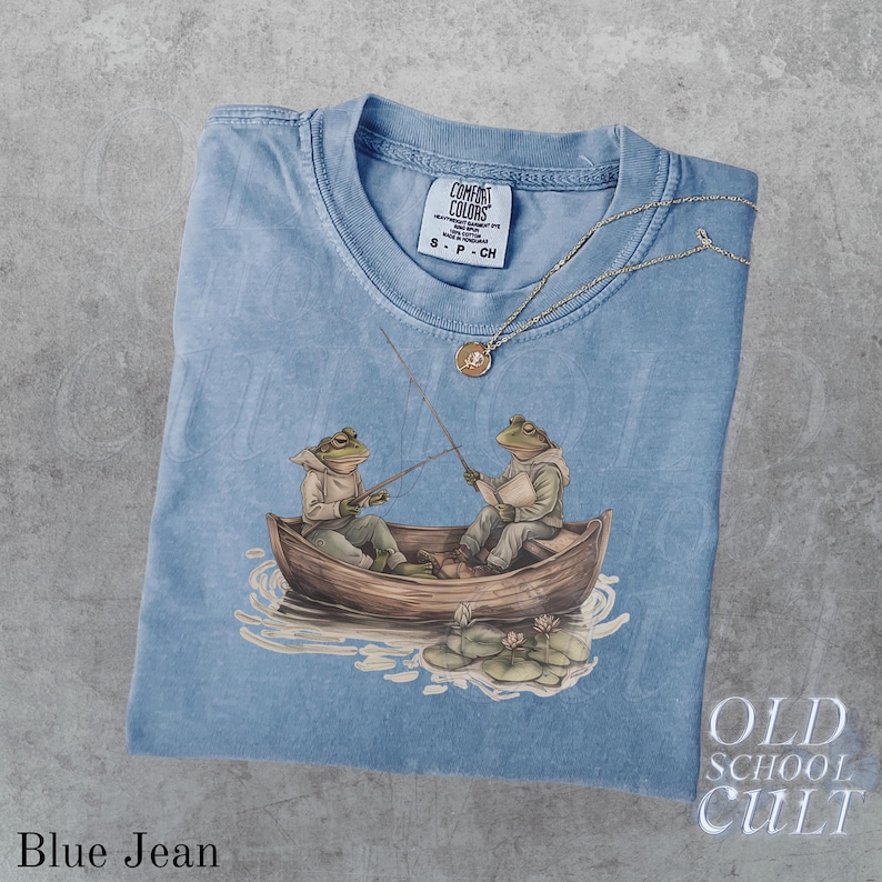 Pode incluir: Camiseta azul jeans com um gr&aacute;fico de dois sapos pescando em um barco. A camisa tem gola redonda e uma pequena etiqueta na gola. Um colar de ouro com um pingente est&aacute; drapeado sobre a camisa. As palavras "OLD SCHOOL CULT" s&atilde;o impressas no canto inferior direito.