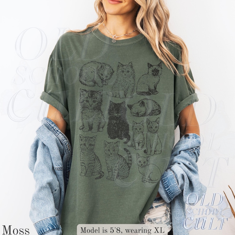 May include: Moss green t-shirt featuring a grid of illustrated cats in various poses. The shirt has a relaxed fit and rolled sleeves. The text "OLD SCHOOL CULT" is visible on the lower right. The model is 5'8" and wearing an XL.