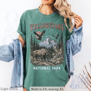May include: Light green t-shirt with a graphic of Yellowstone National Park. The design includes a bison, wolves, eagles, mountains, and trees. The text "YELLOWSTONE NATIONAL PARK" is printed across the top and bottom of the graphic.