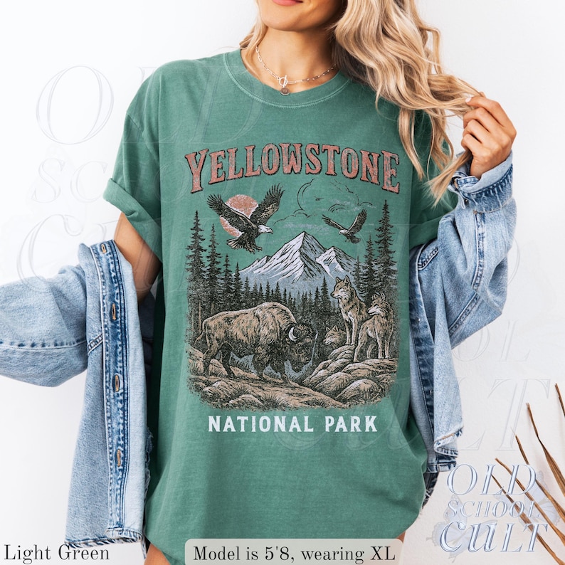May include: Light green t-shirt with a graphic of Yellowstone National Park. The design includes a bison, wolves, eagles, mountains, and trees. The text "YELLOWSTONE NATIONAL PARK" is printed across the top and bottom of the graphic.