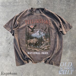 May include: Espresso-colored t-shirt with a graphic of Yellowstone National Park. The design features a bison, wolves, eagles, mountains, and trees. The text "YELLOWSTONE NATIONAL PARK" is printed across the top and bottom of the graphic. The words "OLD SCHOOL CULT" are printed on the bottom right.