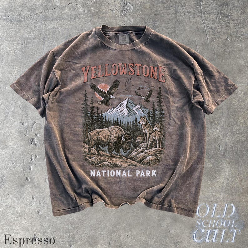 May include: Espresso-colored t-shirt with a graphic of Yellowstone National Park. The design features a bison, wolves, eagles, mountains, and trees. The text "YELLOWSTONE NATIONAL PARK" is printed across the top and bottom of the graphic. The words "OLD SCHOOL CULT" are printed on the bottom right.