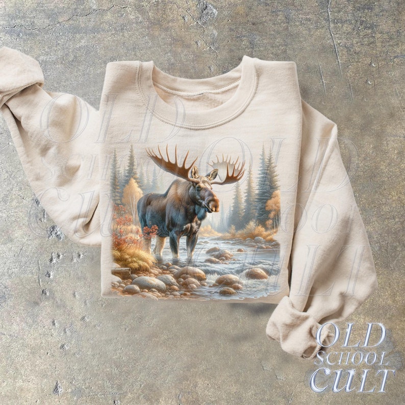 May include: A beige sweatshirt with a graphic print of a moose standing in a stream. The moose is brown with large antlers. The background is a forest scene with trees and rocks.