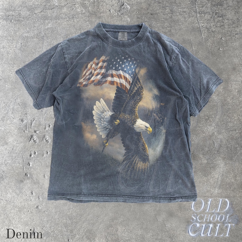 May include: A denim-blue t-shirt with a print of a bald eagle and an American flag. The eagle is in flight, with a misty landscape in the background. The shirt has a faded look. The words "OLD SCHOOL CULT" are printed on the bottom right.
