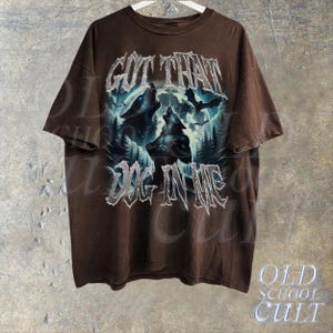 K&ouml;nnte beinhalten: Braunes T-Shirt mit einem grafischen Druck von drei W&ouml;lfen, die zum Mond heulen. Der Text "GOT THAT DOG IN ME" ist in silbernem Glitzer auf der Vorderseite des Shirts gedruckt.