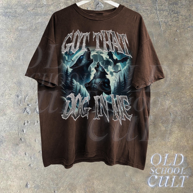 K&ouml;nnte beinhalten: Braunes T-Shirt mit einem grafischen Druck von drei W&ouml;lfen, die zum Mond heulen. Der Text "GOT THAT DOG IN ME" ist in silbernem Glitzer auf der Vorderseite des Shirts gedruckt.