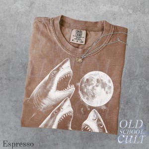 May include: Espresso-colored t-shirt with a graphic of sharks and a full moon. The shirt has a crew neck and short sleeves. A silver necklace with a small pendant rests on the shirt. The text "OLD SCHOOL CULT" is visible.