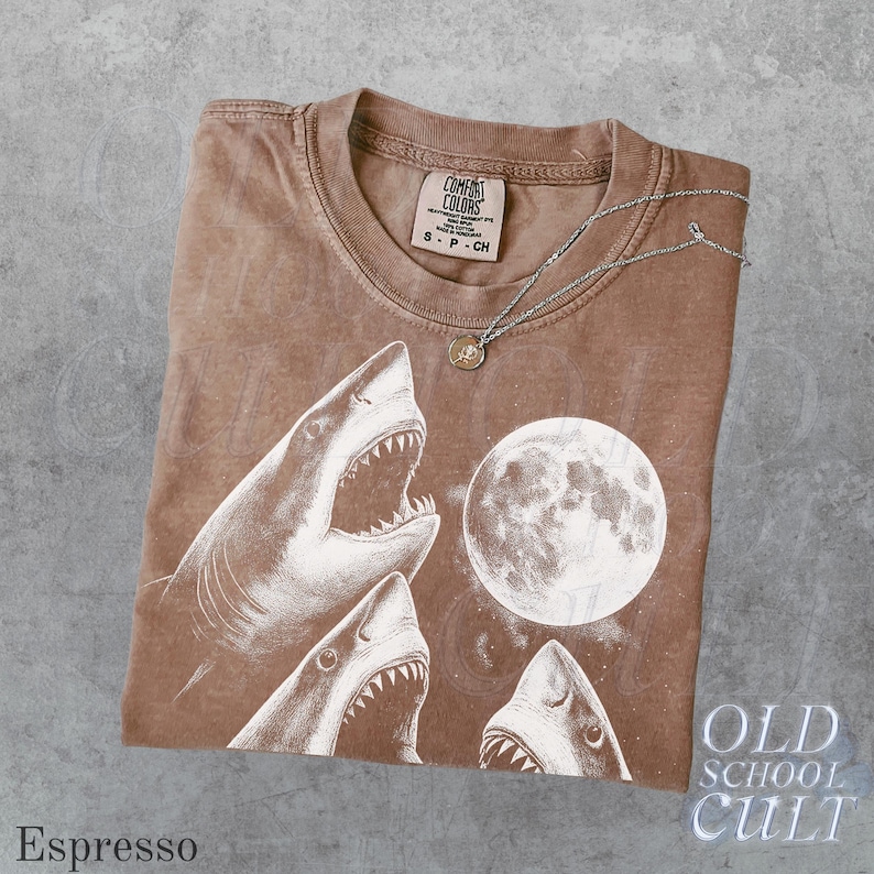 May include: Espresso-colored t-shirt with a graphic of sharks and a full moon. The shirt has a crew neck and short sleeves. A silver necklace with a small pendant rests on the shirt. The text "OLD SCHOOL CULT" is visible.