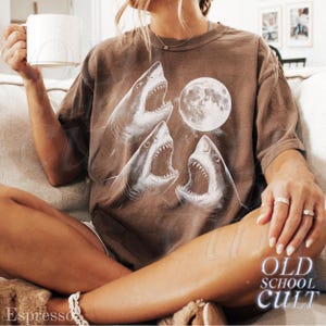 May include: Brown t-shirt featuring a graphic of three open-mouthed sharks and a full moon. The shirt has the text "OLD SCHOOL CULT" in a stylized font. The word "Espresso" is also visible.