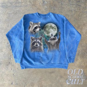 Op de afbeelding: Een blauw sweatshirt met een grafische print van drie wasberen die naar een volle maan kijken. De wasberen zijn in een realistische stijl en de maan is wit met een lichtblauwe gloed.