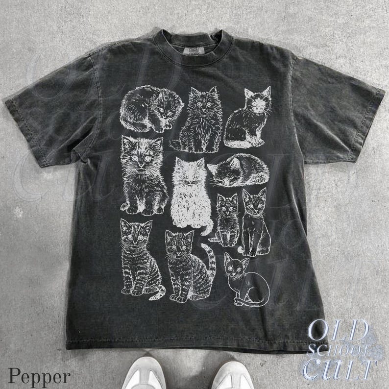 May include: Dark gray t-shirt featuring a collection of illustrated cats in various poses. The shirt has a crew neck and short sleeves. The text "OLD SCHOOL CULT" is printed on the lower right side of the shirt.