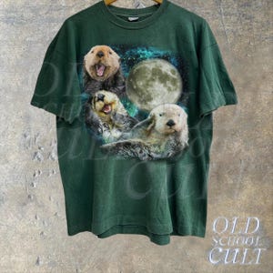 May include: A dark green t-shirt with a graphic print of three sea otters swimming in a starry night sky with a full moon. The otters are white and brown with black eyes.