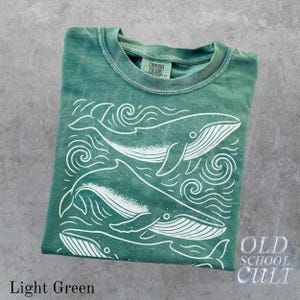 May include: Light green t-shirt with a white whale graphic and the text "OLD SCHOOL CULT". The shirt features a crew neck and short sleeves. The graphic includes three whales and wave designs.