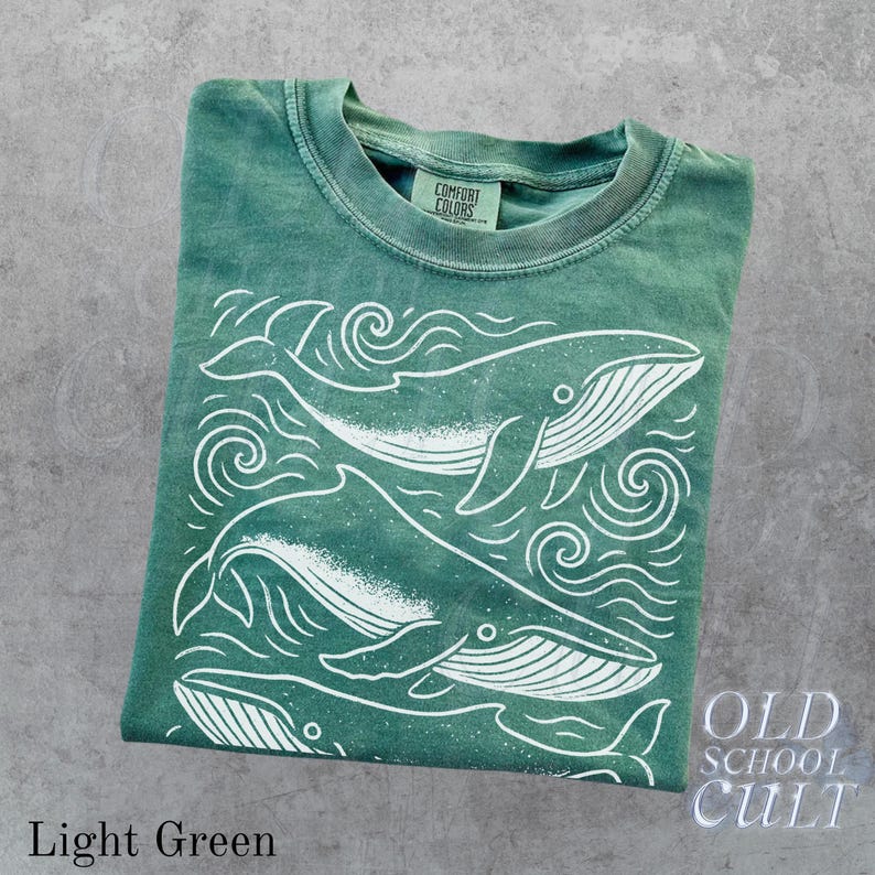 May include: Light green t-shirt with a white whale graphic and the text "OLD SCHOOL CULT". The shirt features a crew neck and short sleeves. The graphic includes three whales and wave designs.