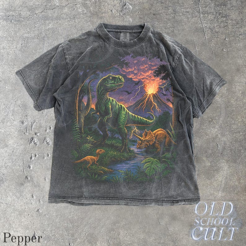 Op de afbeelding: Donkergrijze t-shirt met een kleurrijke dinosaurusillustratie, waaronder een groene Tyrannosaurus Rex, Triceratops en een vulkaanuitbarsting tegen een zonsondergang. De t-shirt heeft een vintage uitstraling. De tekst "OLD SCHOOL CULT" is zichtbaar.