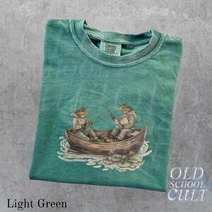 Pode incluir: Camiseta verde claro com um gr&aacute;fico de dois sapos pescando em um barco. Os sapos usam jaquetas com capuz e seguram varas de pesca. O texto "OLD SCHOOL CULT" est&aacute; no canto inferior direito.